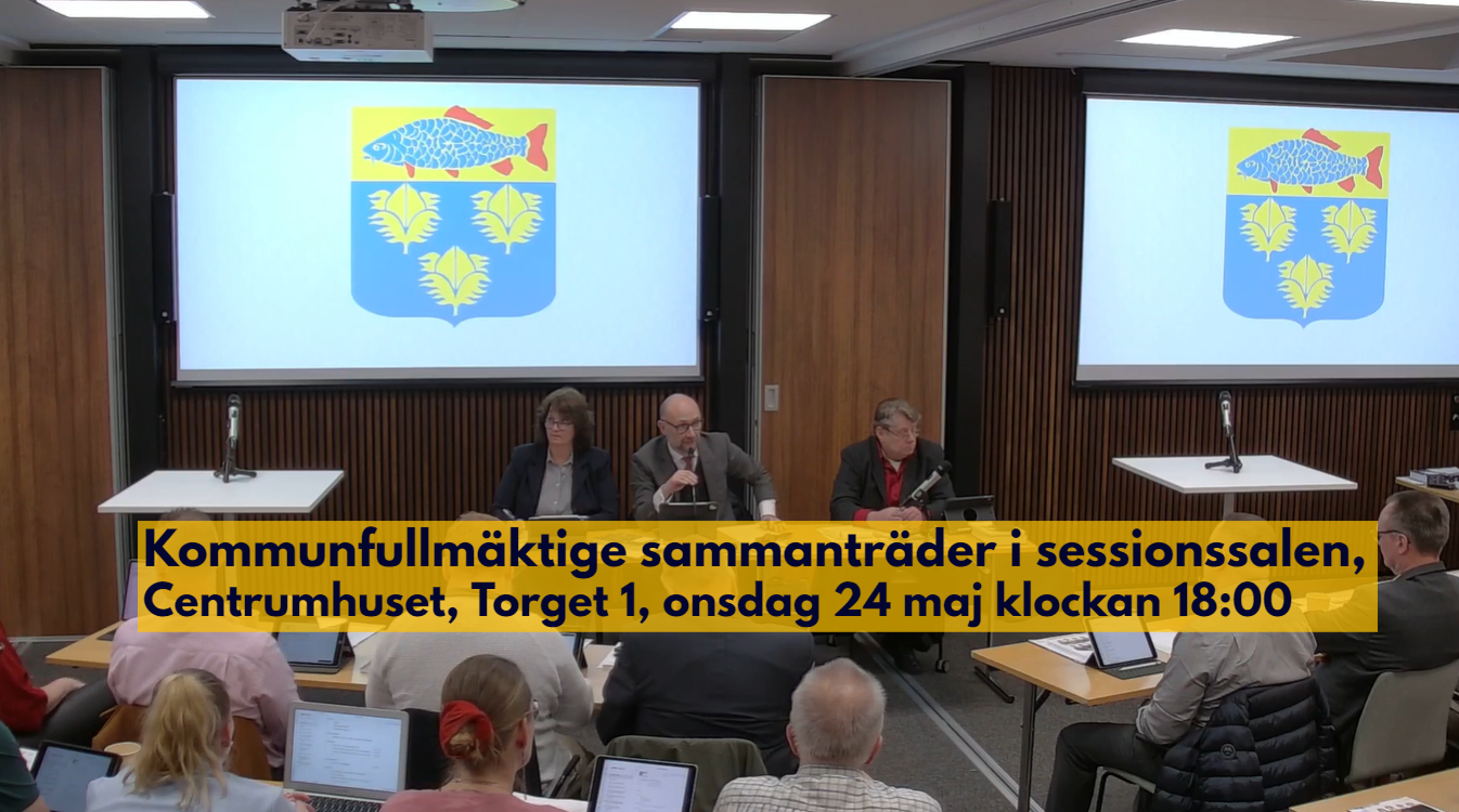 Kommunfullmäktige sammanträder i sessionssalen, Centrumhuset, onsdag 24 maj 2023. Informations står med mörk text på gul bakgrund över bilden när man ser kommunfullmäktiges presidie och kommunens vapen på skärmar bakom..