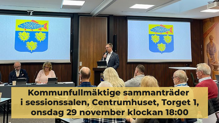 Kommunfullmäktige sammanträder i sessionssalen, Centrumhuset, onsdag 29 november 2023. Information står med mörk text på beige bakgrund över bilden där man ser kommunfullmäktiges presidie och kommunens vapen på skärmar bakom.