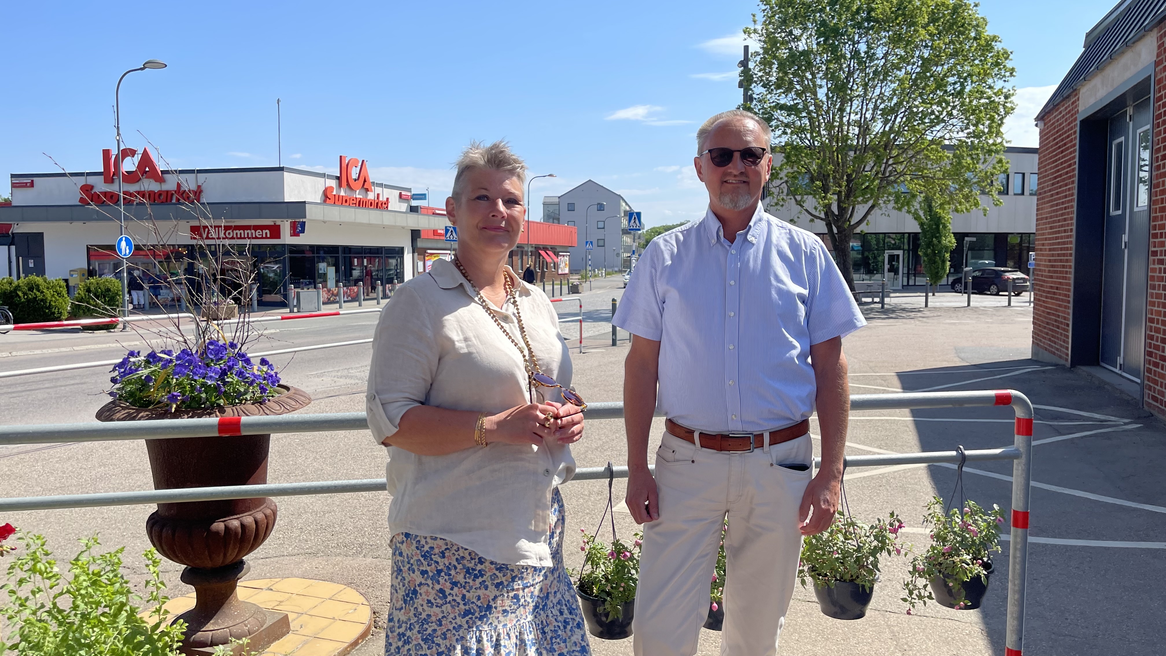 En man och en kvinna står bredvid varandra bredvid en blomsterurna. I bakgrunden ses ICA:s entré och Centrumhuset i Perstorp.