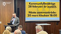 Bild från kommunfullmäktige med skärm på vägg i bakgrunden på vilken det står skrivet med svart text på gul bakgrund att kommunfullmäktige den 28 februari är inställd och att nästa sammanträde är den 20 mars.