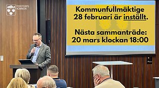 Bild från kommunfullmäktige med skärm på vägg i bakgrunden på vilken det står skrivet med svart text på gul bakgrund att kommunfullmäktige den 28 februari är inställd och att nästa sammanträde är den 20 mars.