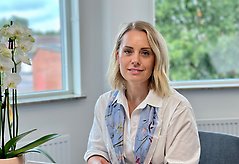 Kvinna sitter framför två fönster och tittar in i kameran. Hon har ljust hår och vit skjorta.