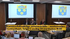 Kommunfullmäktige sammanträder i sessionssalen, Centrumhuset, onsdag 24 maj 2023. Informations står med mörk text på gul bakgrund över bilden när man ser kommunfullmäktiges presidie och kommunens vapen på skärmar bakom..