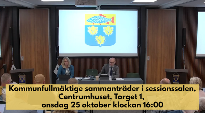 Kommunfullmäktige sammanträder i sessionssalen, Centrumhuset, onsdag 25 oktober 2023. Information står med mörk text på beige bakgrund över bilden där man ser kommunfullmäktiges presidie och kommunens vapen på skärmar bakom.