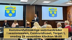 Kommunfullmäktige sammanträder i sessionssalen, Centrumhuset, onsdag 29 november 2023. Information står med mörk text på beige bakgrund över bilden där man ser kommunfullmäktiges presidie och kommunens vapen på skärmar bakom.