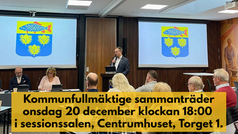 Kommunfullmäktige sammanträder i sessionssalen, Centrumhuset, onsdag 20 december 2023. Information står med mörk text på gul bakgrund över bilden där man ser kommunfullmäktiges presidie och kommunens vapen på skärmar bakom.