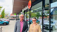 En man och en kvinna står på en trottoar utanför buitiker. i bakgrunden ses skyltfönster och parkerade bilar.